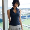テニスコートを望む窓辺に立つネイビーのノースリーブポロと白いプリーツスカート姿の女性